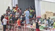 Grupa młodych migrantów w obozie tymczasowym w Ceucie przy barierkach odgradzających, obsługa w rękawiczkach i maseczkach wydaje zaopatrzenie
