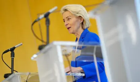 Szefowa Komisji Europejskiej Ursula von der Leyen w niebieskim żakiecie przemawia zza przezroczystej mównicy na żółtym tle.