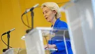Szefowa Komisji Europejskiej Ursula von der Leyen w niebieskim żakiecie przemawia zza przezroczystej mównicy na żółtym tle.